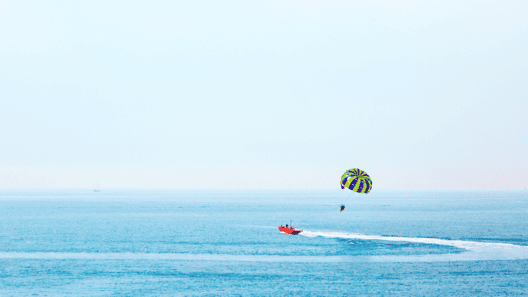 青い海と青空の地平線が広がる中に赤いボートでカラフルなパラシュートを広げてパラセーリングをしている画像