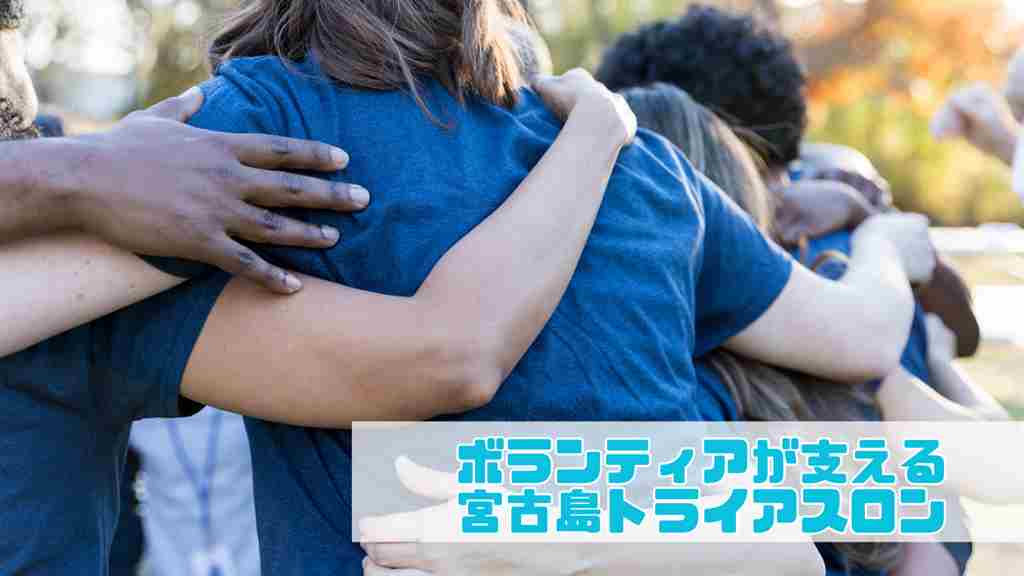 右下に”ボランティアが支える宮古島トライアスロン”と記載。屋外の樹木がある広間を背景にTシャツを着た男女が肩を組み合っている背中を写した画像